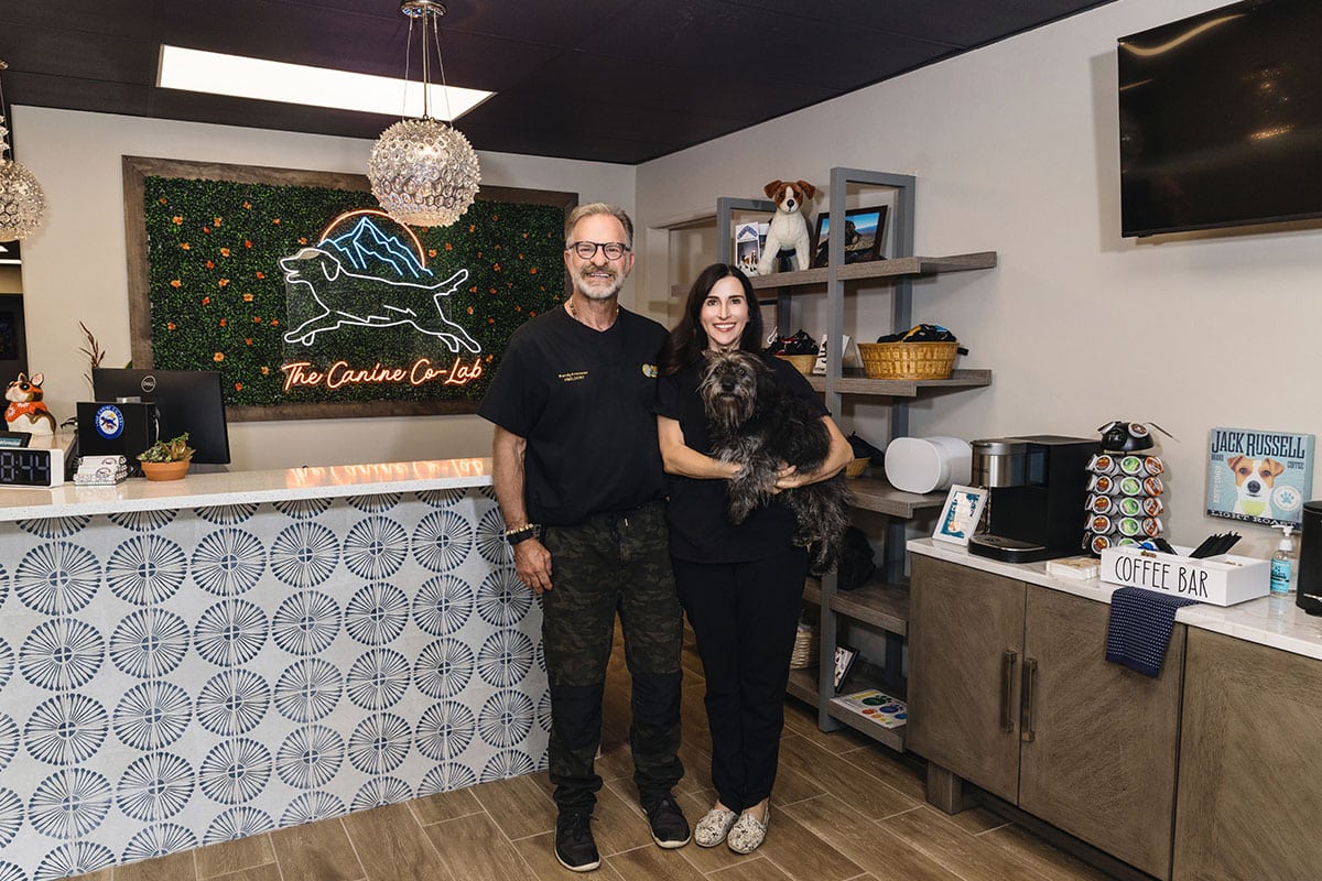 A man and a woman stand in a pet clinic lobby, where the woman holds a small dog. The reception desk boasts a decorative sign with a running dog, while shelves display pet items. A vet-themed coffee bar is on the right, adding charm to this welcoming veterinarian setting.