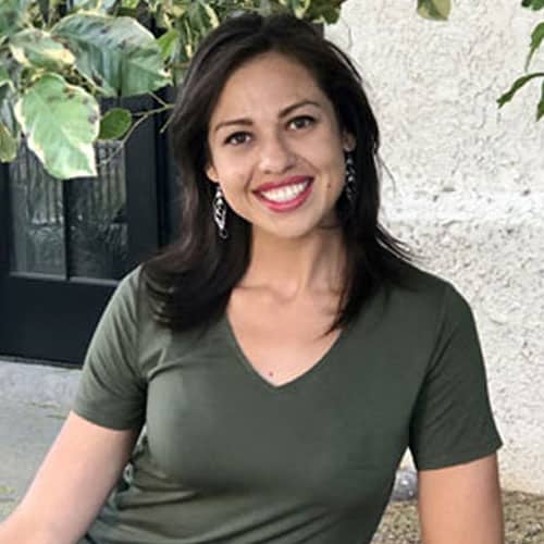 A smiling woman with long dark hair, wearing a green shirt, sits on her porch. As a dedicated veterinarian, she finds solace in the peaceful setting of green leaves and a white wall in the background.