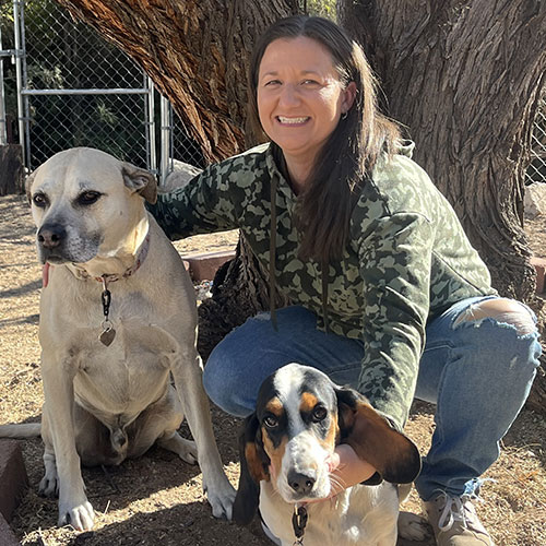 Smiling person in a green camo hoodie kneels outside next to two dogs under a tree, exuding the warmth of a dedicated veterinarian. One dog is large and pale, sitting upright, while the other, smaller with long ears and tricolor fur, rests close by. A fenced area can be seen in the background.