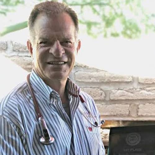 A smiling veterinarian in a striped shirt with a stethoscope around his neck stands in front of a light brick wall, with greenery visible in the background.