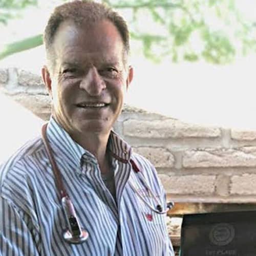 A smiling veterinarian with a short haircut, wearing a striped shirt and a stethoscope around his neck, stands outdoors. A blurred background of leafy greenery and a brick wall is visible.